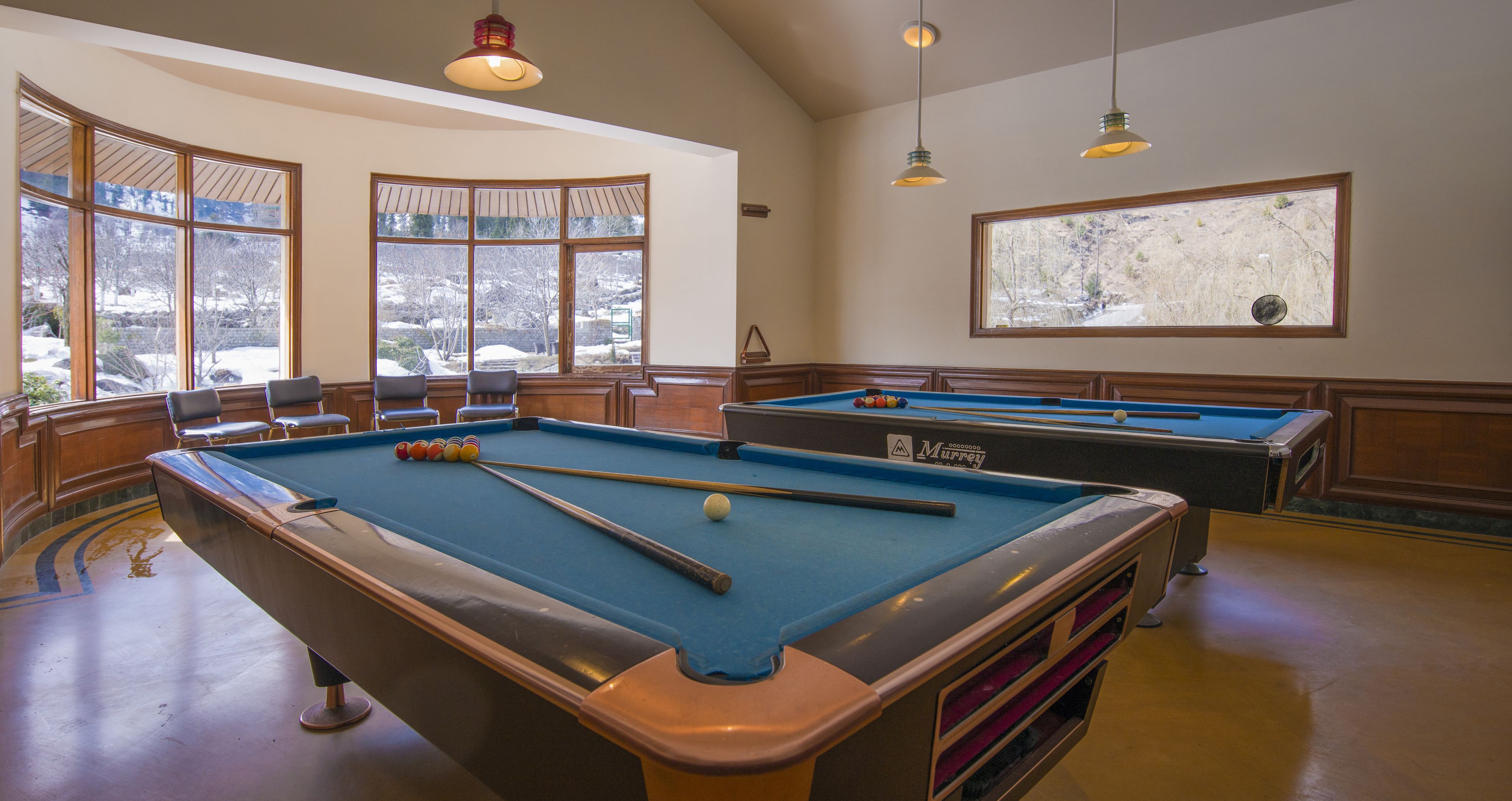 A pool table with a blue felt surface, a pool cue, and several billiard balls. There are two windows with wooden frames, and a large window with a view of trees and snow.