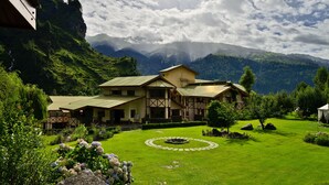 A large, two-story building with a traditional architectural style, surrounded by lush greenery and mountains in the background. - Solang Valley Resort (Manali)