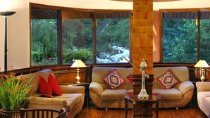 A cozy living room with a fireplace, comfortable seating, and large windows offering a view of a lush garden. - Solang Valley Resort (Manali)