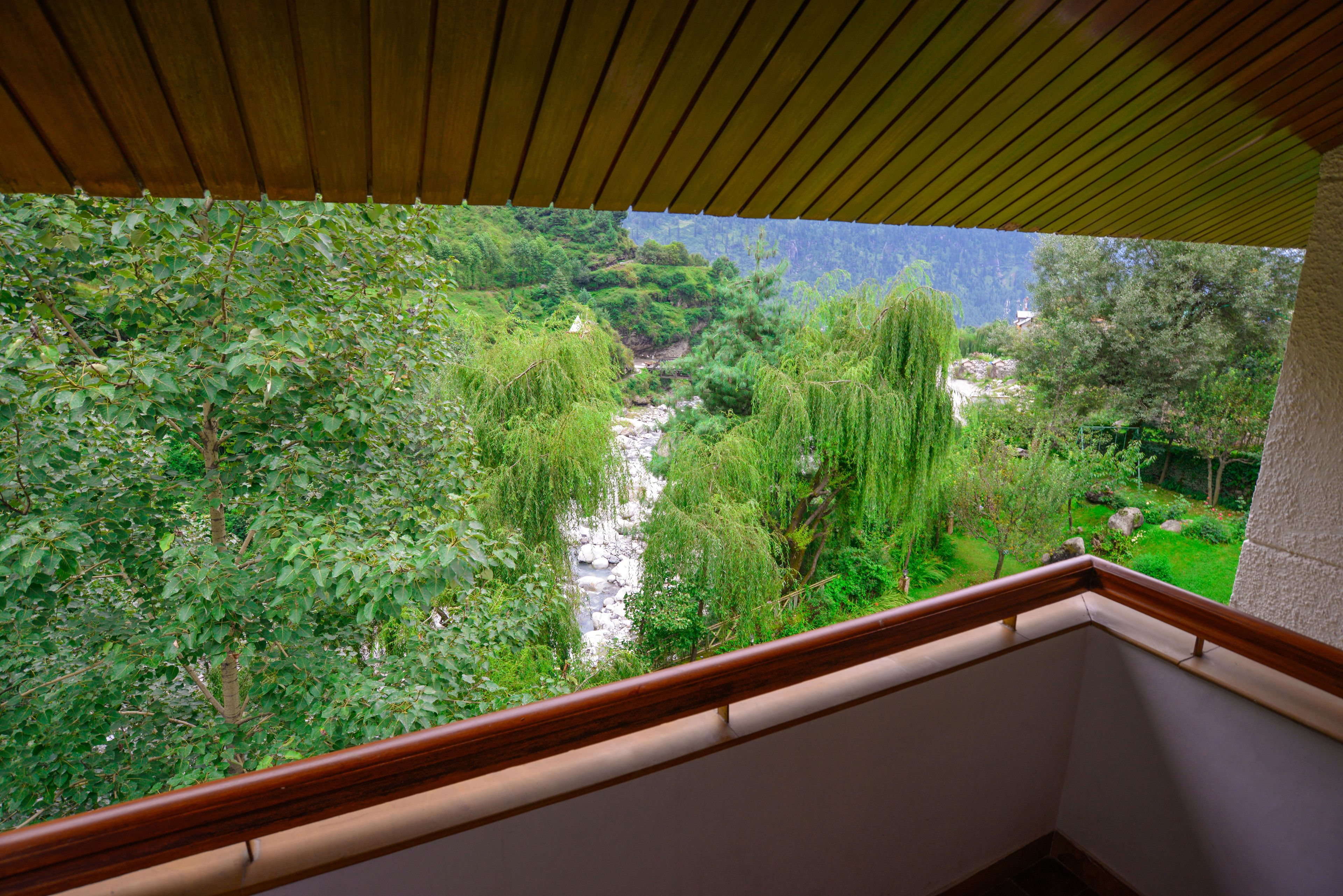 A view from a balcony with a wooden railing, overlooking a lush garden with trees and a stream.