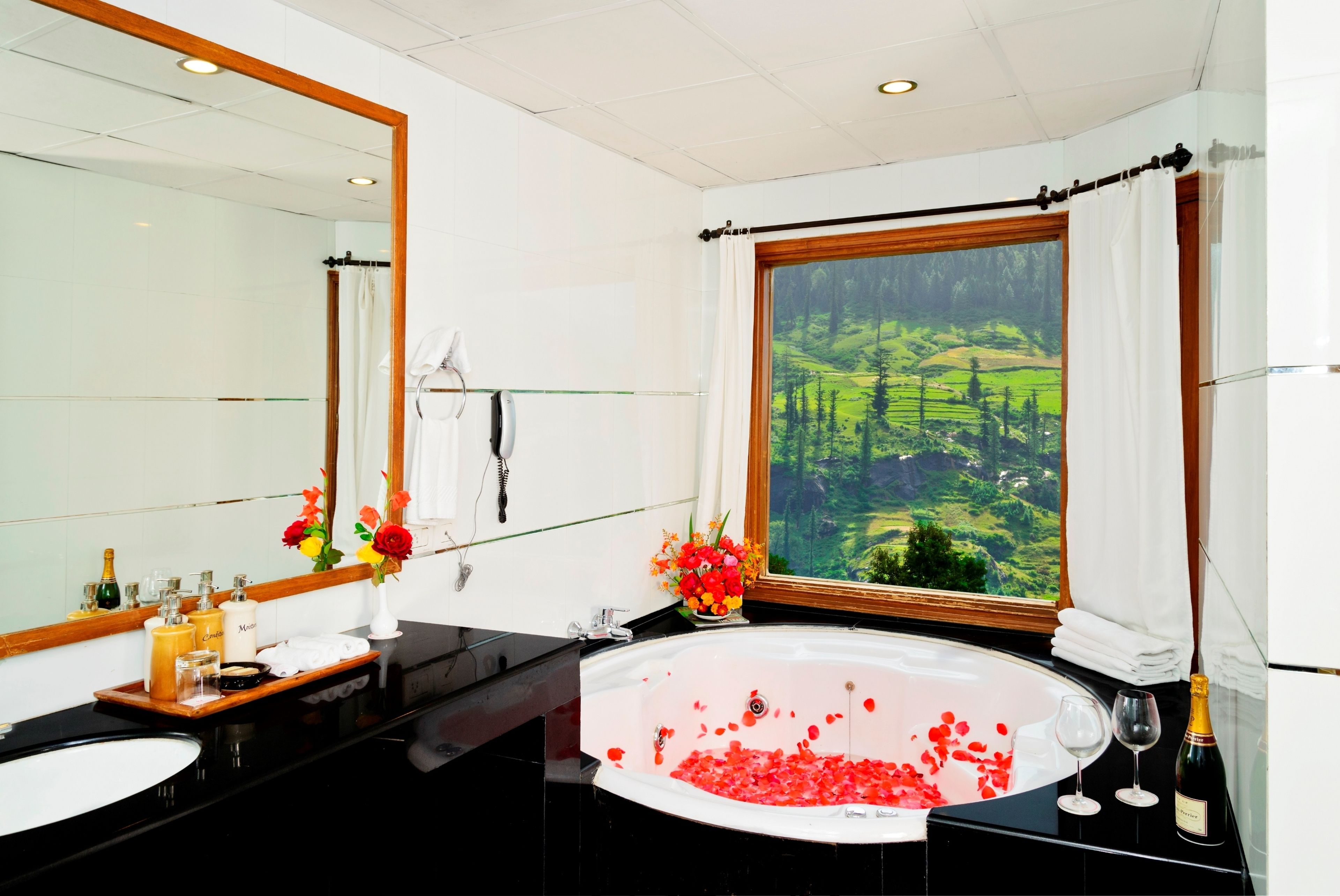 Een badkamer met een groot bad gevuld met rozenblaadjes, een wastafel, een badkamermeubel met toiletartikelen en een raam met een prachtig uitzicht.