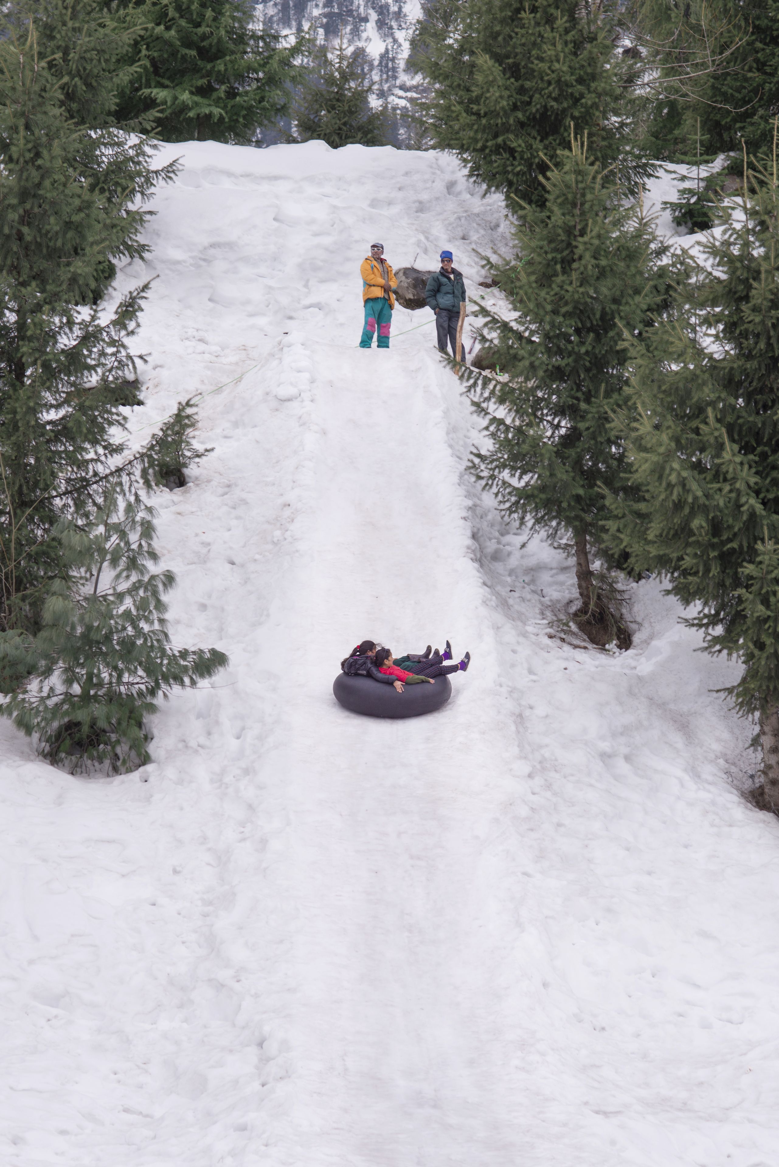 A snowy slope with a person in a yellow jacket and another in a blue jacket standing at the top, and a person sliding down a snow tube.