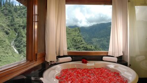 A bathroom with a bathtub filled with rose petals, a window with curtains, and a view of a mountainous landscape. - Solang Valley Resort (Manali)