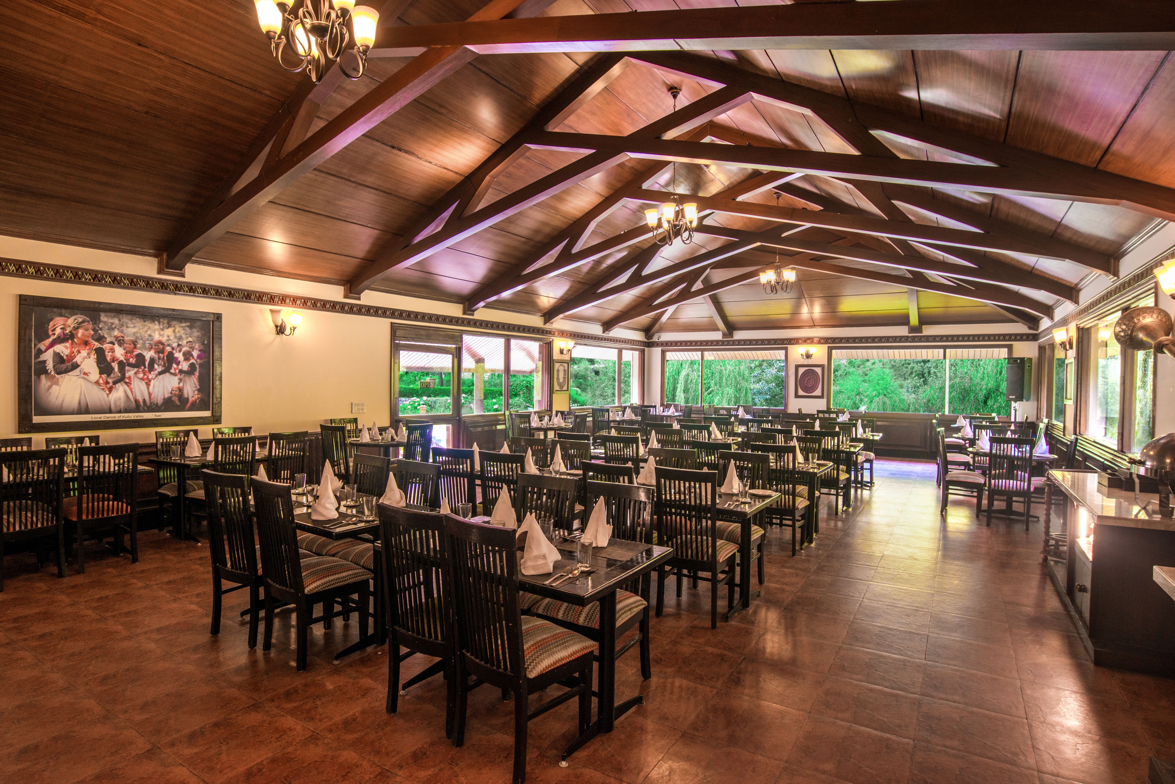 A spacious dining area with wooden tables and chairs, large windows with a view of greenery, and a high ceiling with exposed beams.