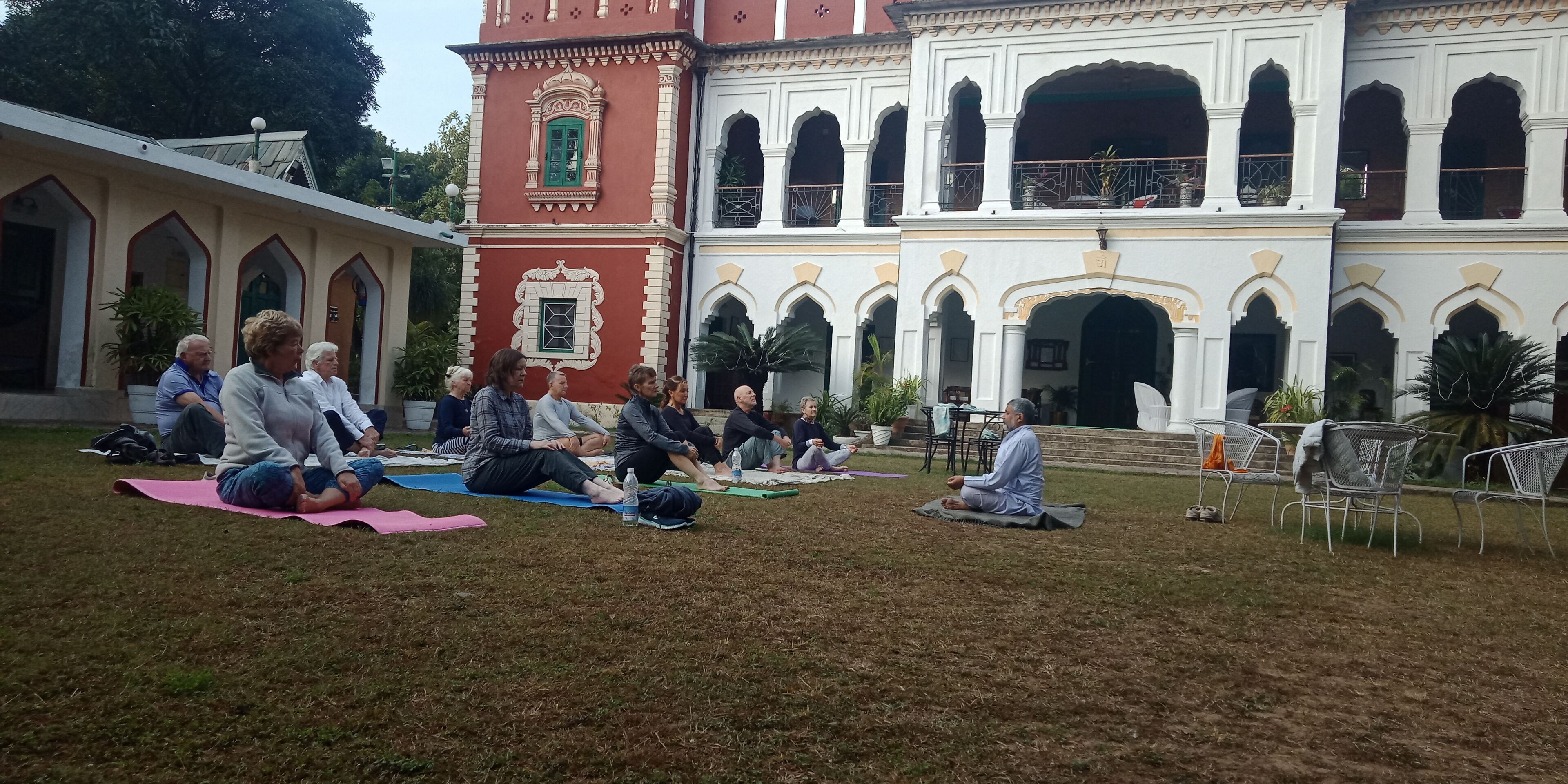 A courtyard with people sitting on mats, a building with arches, and a red tower.