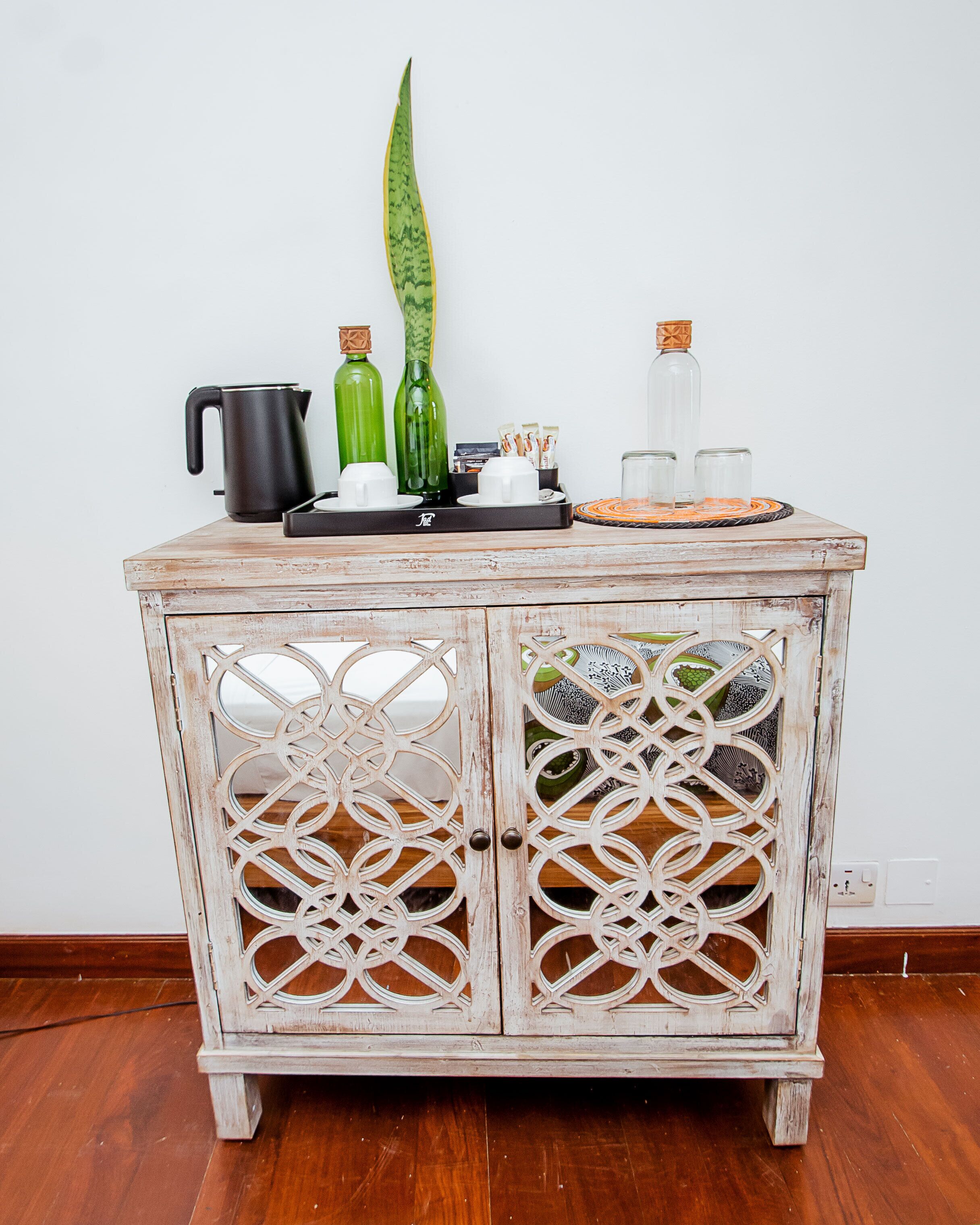 Un meuble en bois orné de motifs complexes découpés dans les portes, un plateau avec des bouteilles en verre et une bouilloire noire posée dessus, et un set de table en fibres tressées.