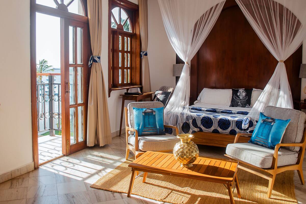 Una habitación con cama, una mesa de madera y una silla. Hay una ventana con cortinas y vistas al exterior.