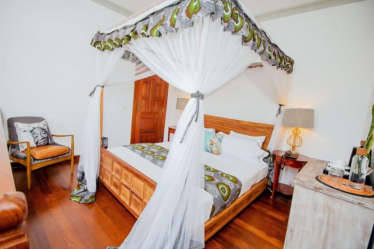 Une chambre à coucher avec un lit en bois, un lit à baldaquin, une chaise avec un coussin, une table de chevet avec une lampe et une petite table sur laquelle est posé un objet décoratif.