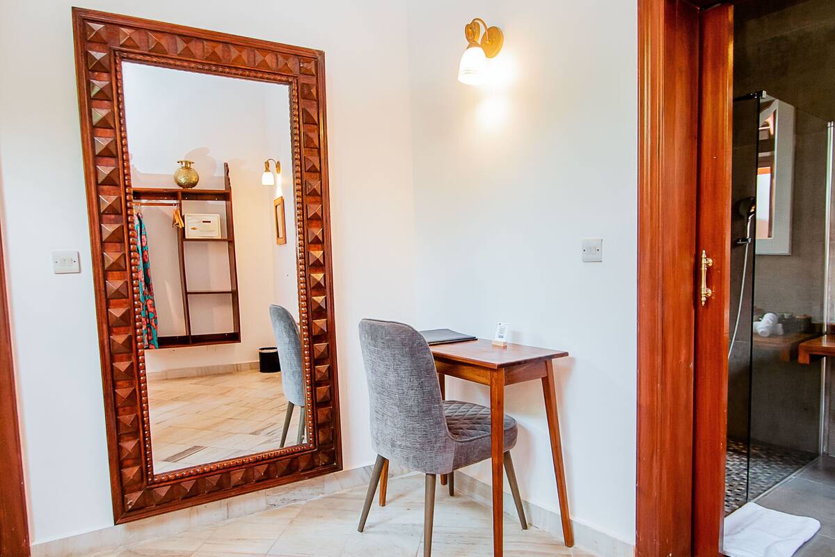 Una habitación con un espejo grande, una mesa de madera, una silla gris y una lámpara fijada a la pared.