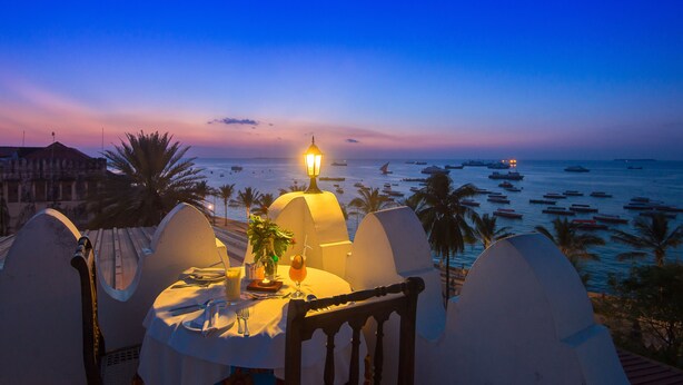 A dining table with a lamp, chairs, and a view of the ocean at dusk.