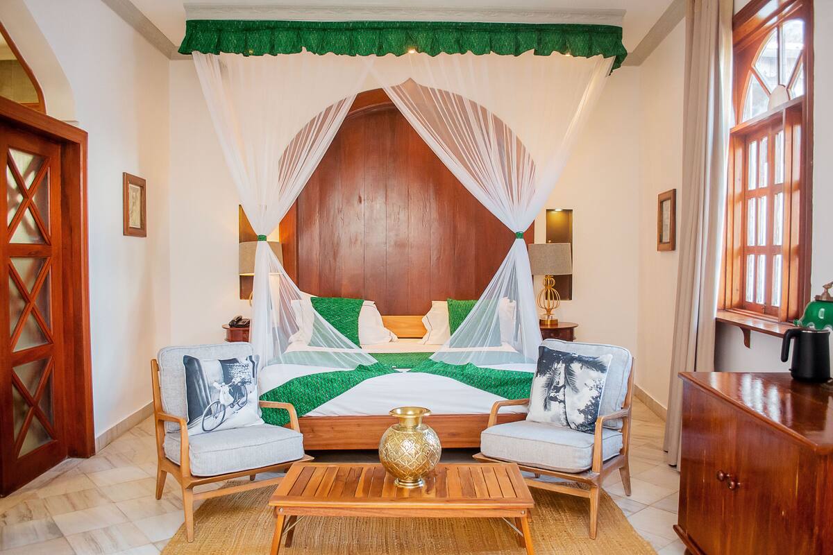 Un dormitorio con cama de madera, dosel, dos sillas, mesita de noche y mesita auxiliar.
