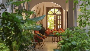 A narrow pathway leading to a seating area with a bench and chairs, surrounded by lush greenery and a large arched window.