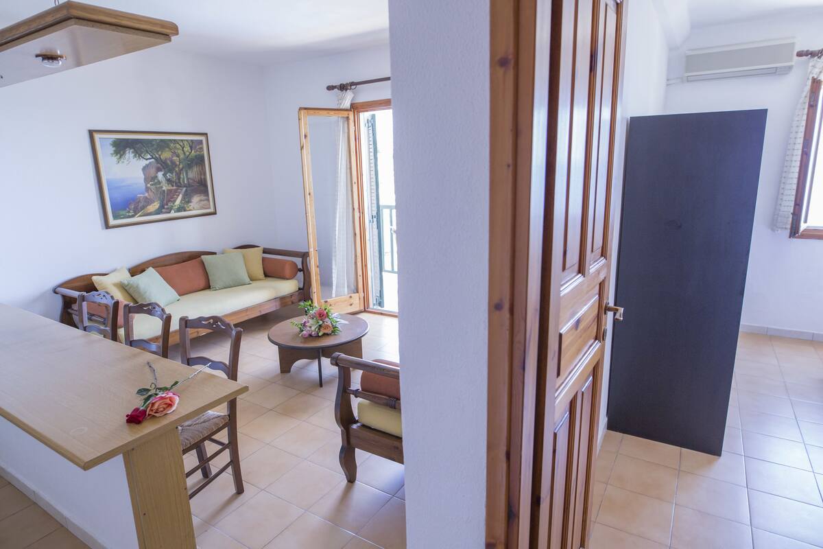 A room with a wooden dining table, a sofa with cushions, a small round table with a flower, and a painting on the wall.