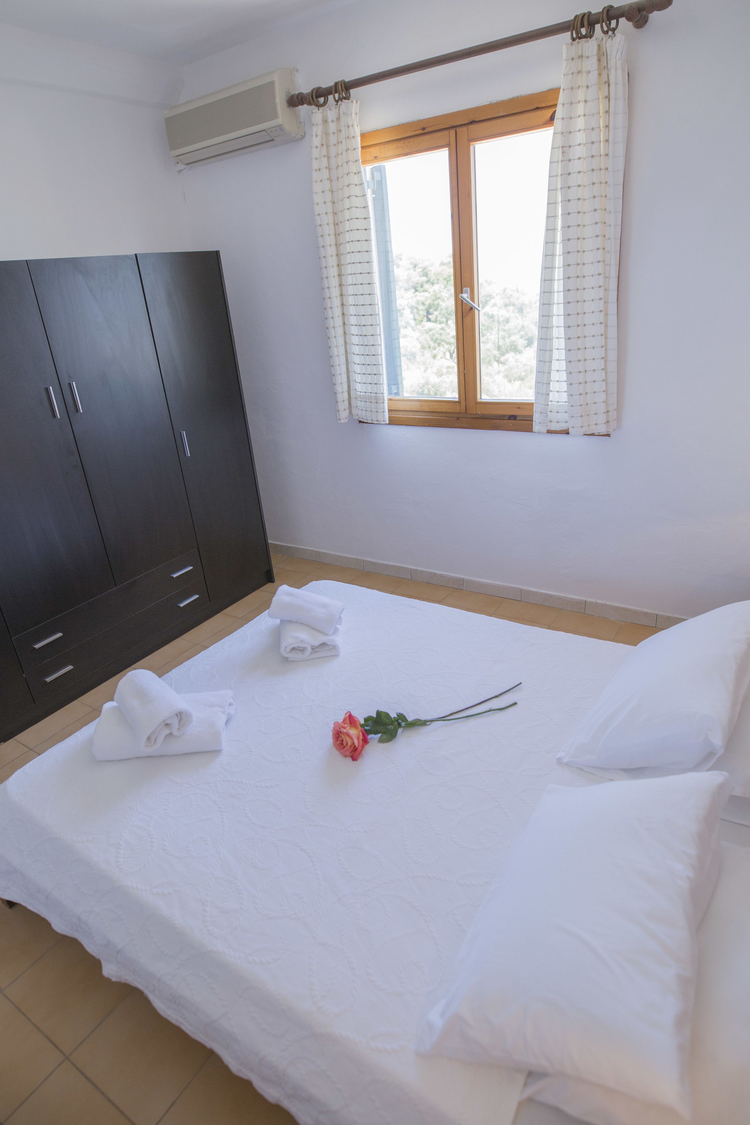 A neatly made bed with white linens and a single rose, a dark wooden wardrobe, and a window with curtains.