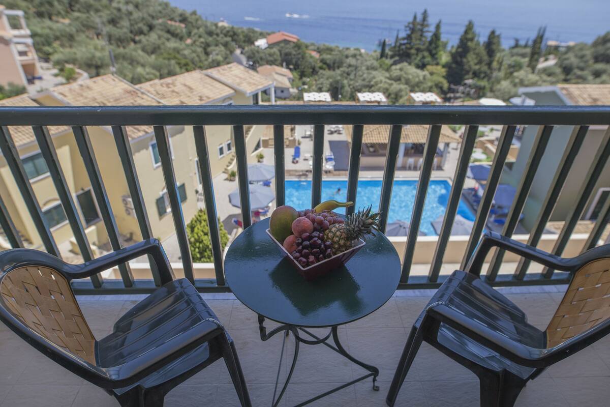 A balcony with a table holding a fruit bowl, two chairs, and a view of a swimming pool and buildings.