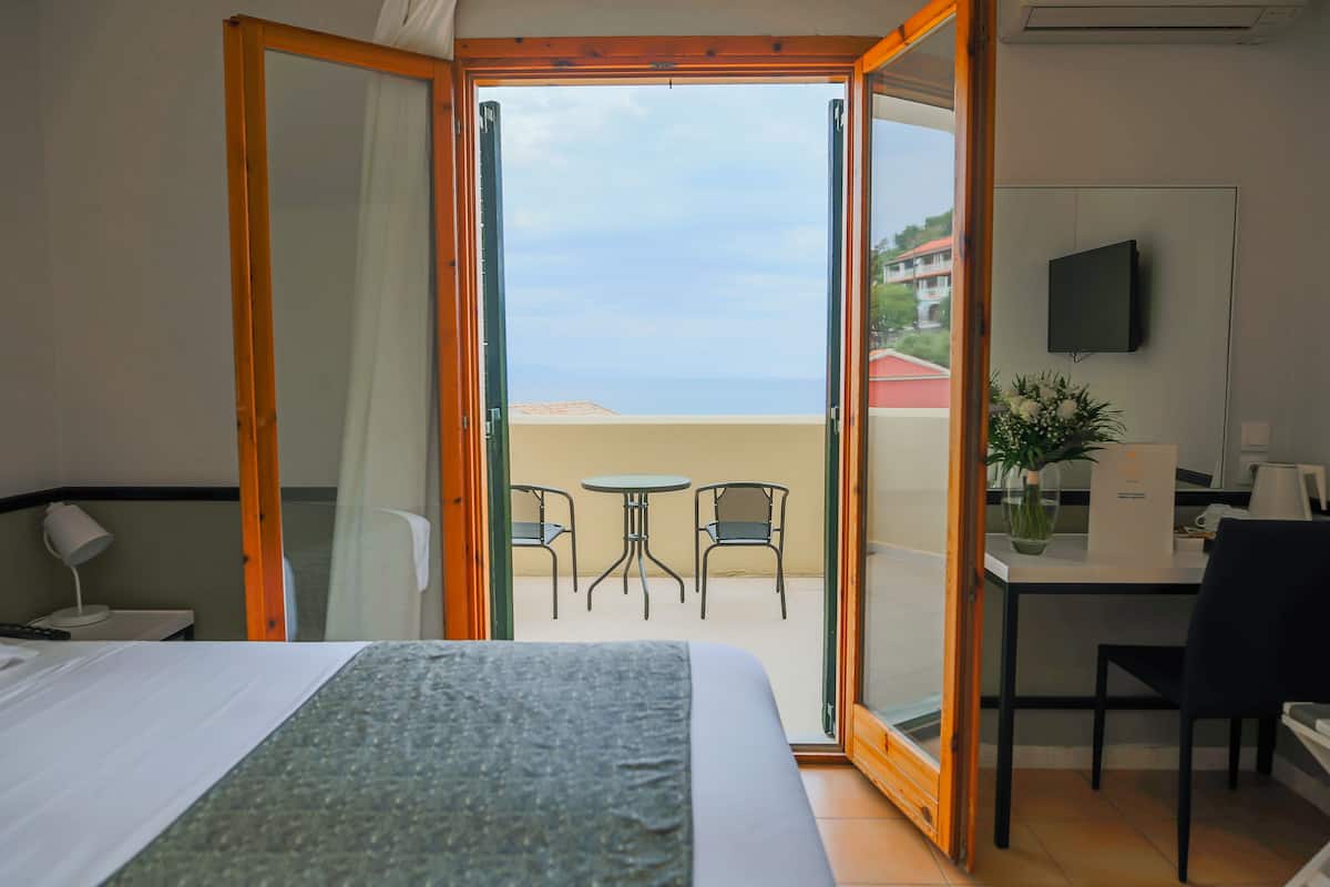 A hotel room with a bed, a desk, a chair, a TV, and a balcony with a view of the sea.