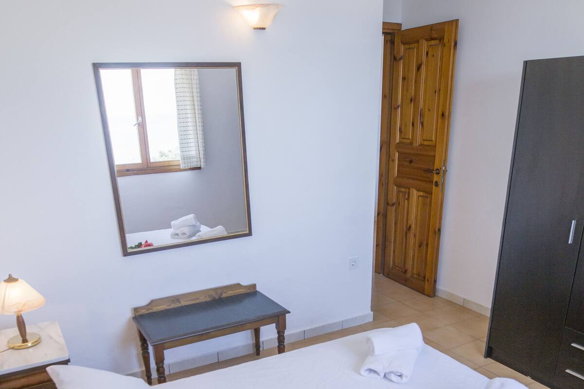 A neatly made bed with white linens, a bedside lamp, a small wooden bench, a mirror on the wall, and a wooden wardrobe.