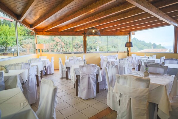 Un restaurante con vigas de madera, manteles blancos y sillas, con un fondo de vegetación y cielo despejado.