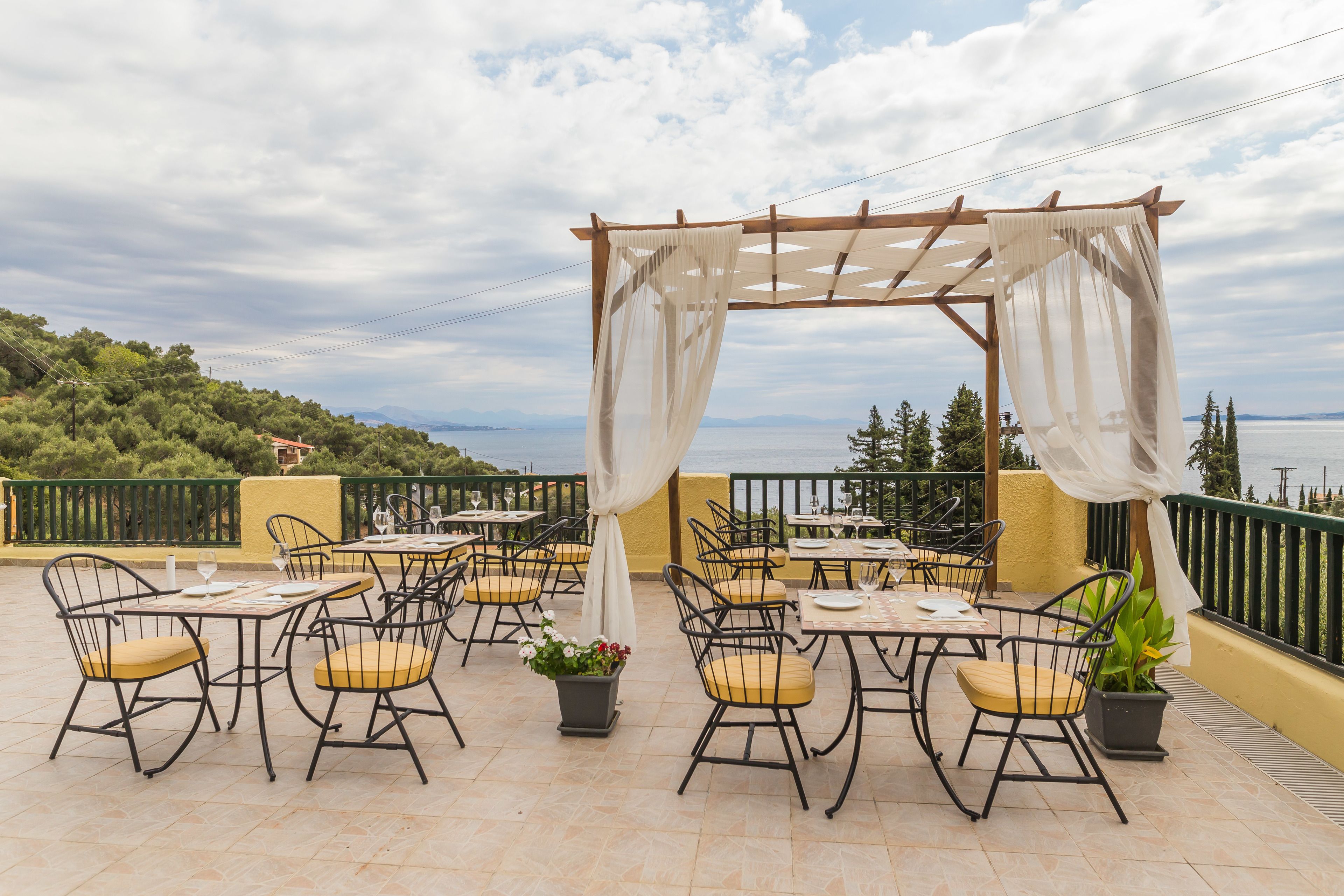 Un patio al aire libre con mesas y sillas, una pérgola con cortinas blancas, y vistas al mar y las colinas.