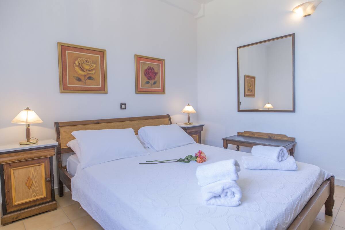 A neatly made bed with white linens, two framed floral paintings on the wall, a bedside table with a lamp, and a mirror.