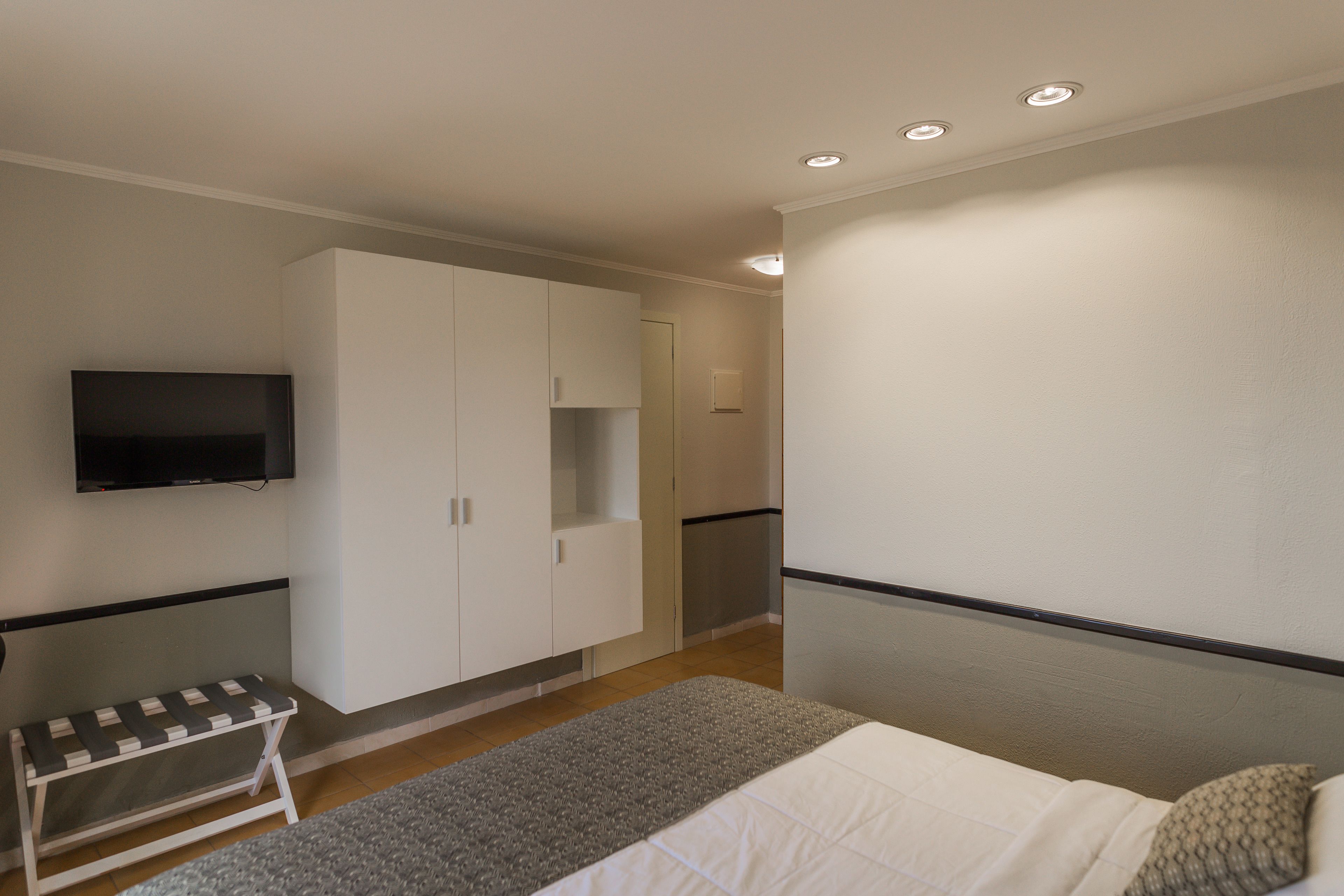 A bedroom with a bed, a bench, a wardrobe, a television, and a small kitchenette.