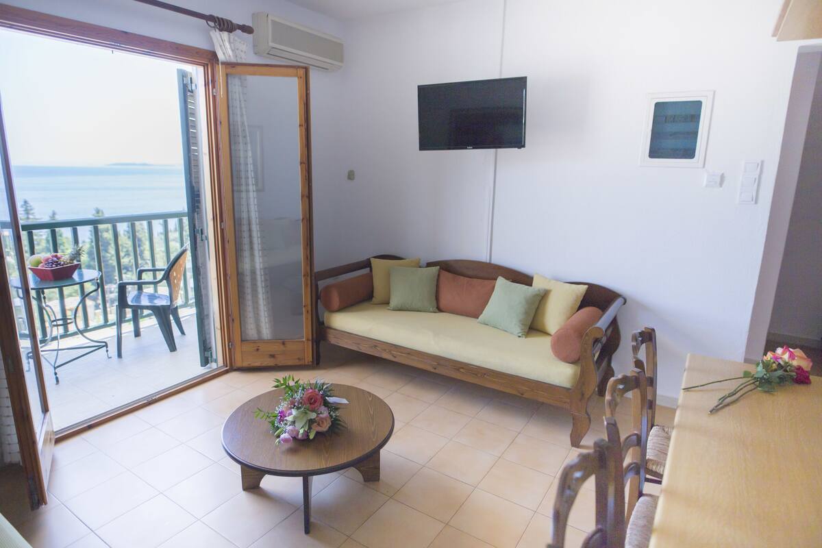 A living room with a sofa, a coffee table with flowers, a TV, and a balcony with a table and chairs.