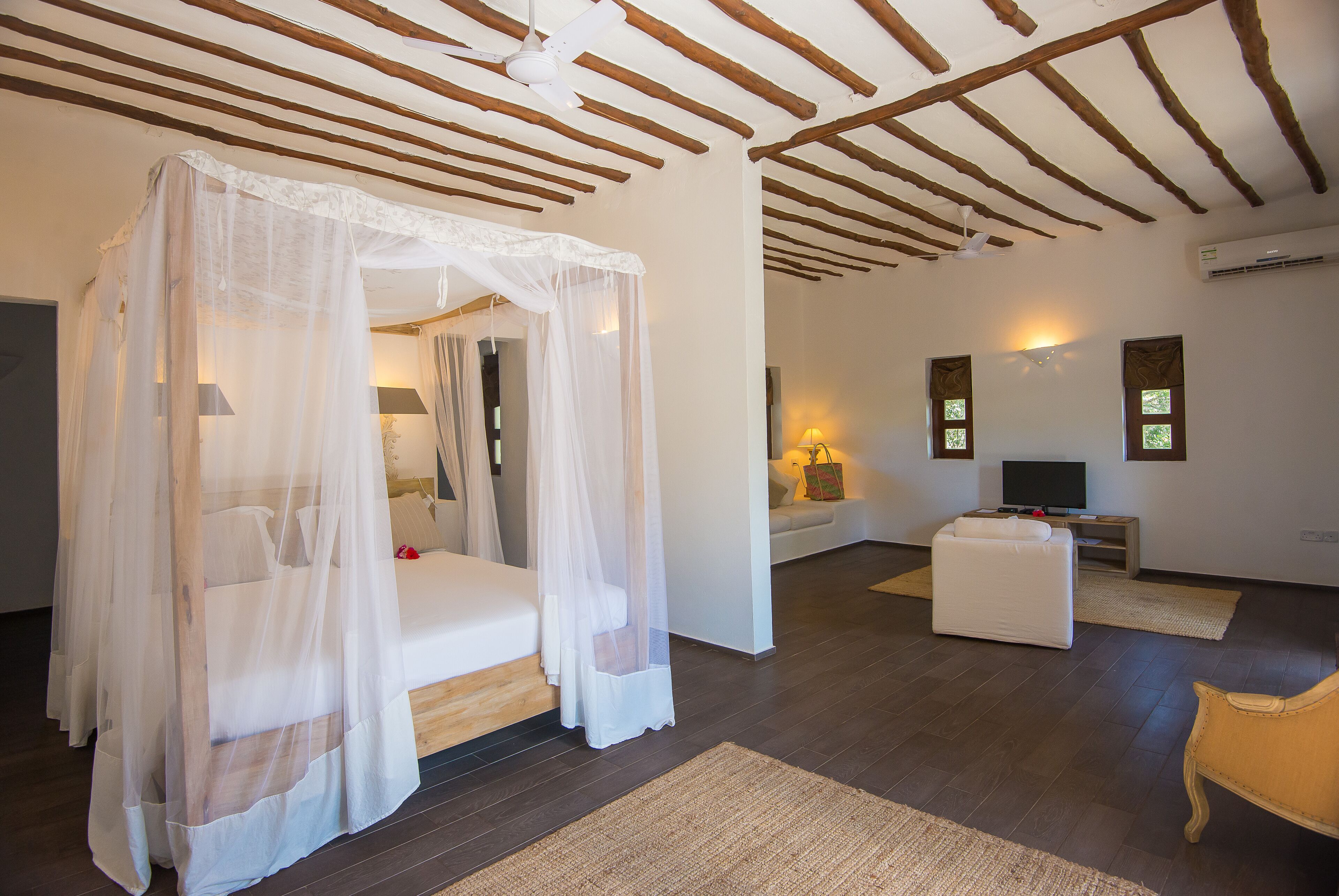 A bedroom with a four-poster bed, a sitting area with a sofa and a TV, and a wooden ceiling.