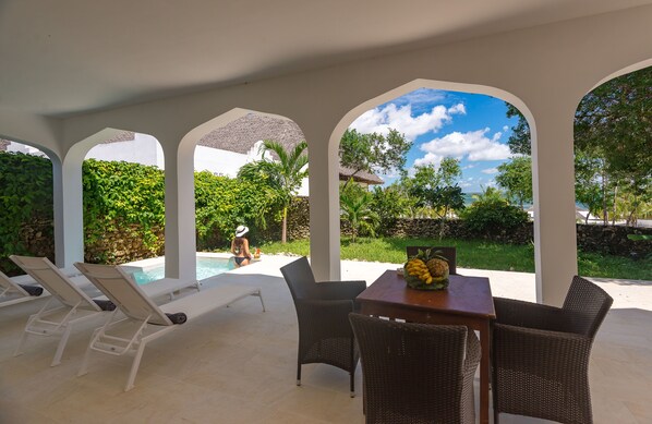 A covered outdoor area with lounge chairs, a table with fruit, and a view of a swimming pool and greenery.