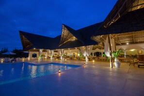 A swimming pool area with a thatched roof structure, lounge chairs, and ambient lighting.