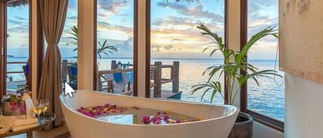 Una bañera grande llena de agua y flores, ubicada cerca de amplios ventanales con vistas al mar.