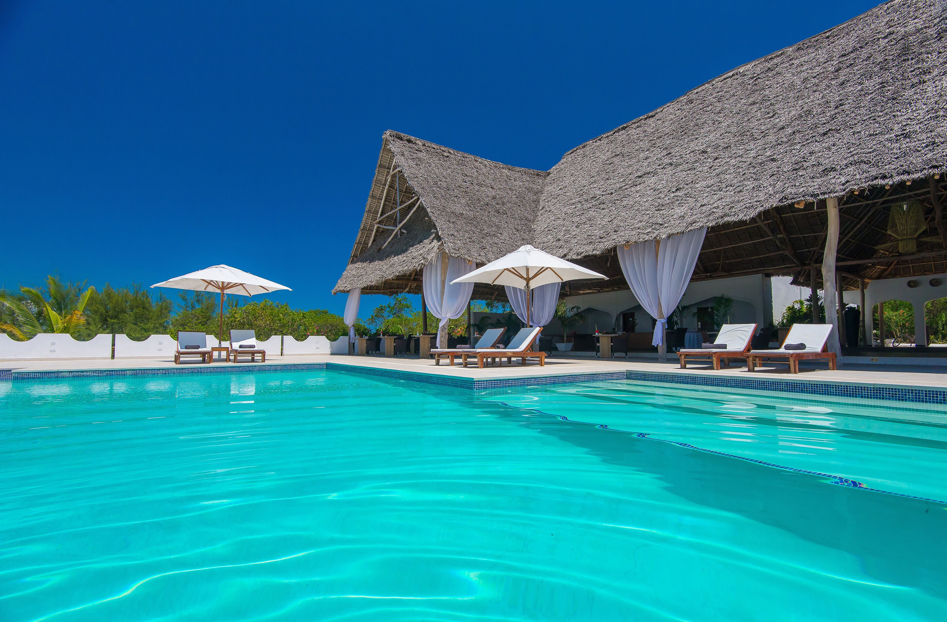 A swimming pool with lounge chairs and umbrellas, surrounded by a thatched-roof structure.
