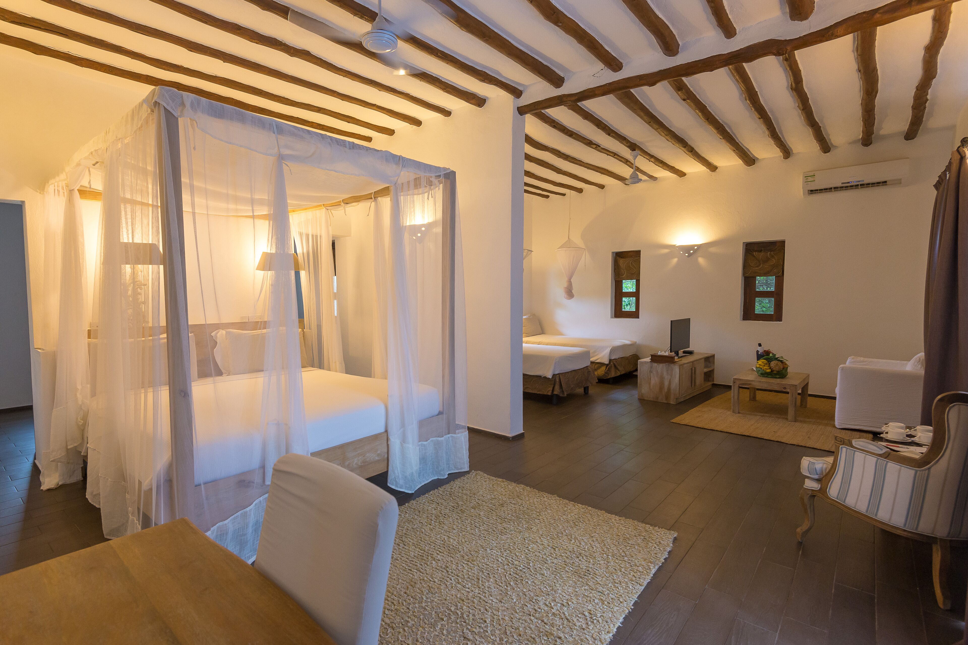 A room with a four-poster bed, a sitting area with a sofa and armchairs, a small table, and a wooden floor.