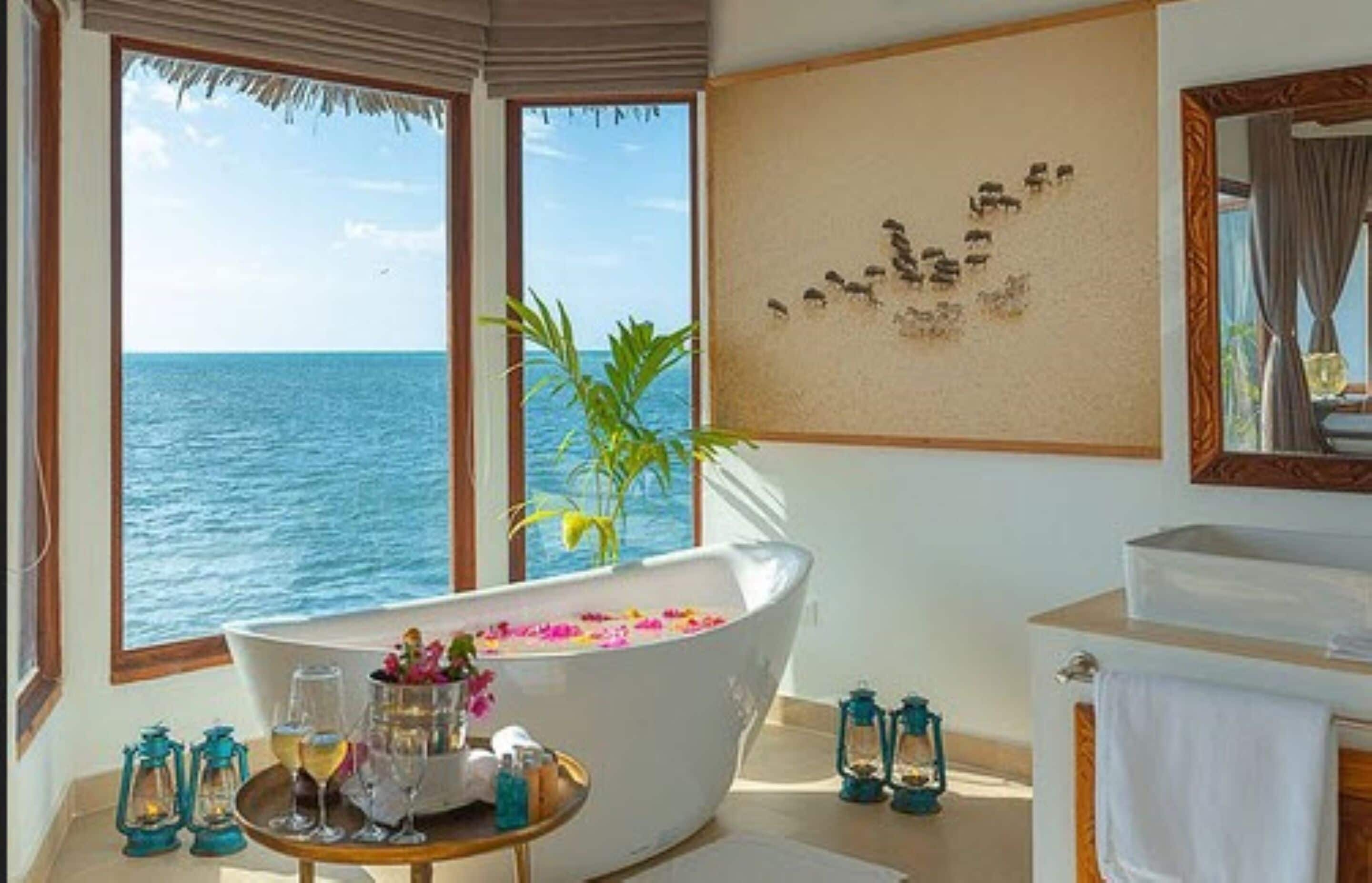 A room with a bathtub filled with flowers, a large window with a view of the sea, a mirror, and a small table with drinks and lanterns.