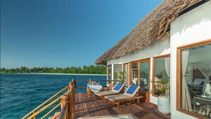 Una terraza de madera con piscina, sillas de descanso y una estructura de techo de paja con vistas al mar turquesa.