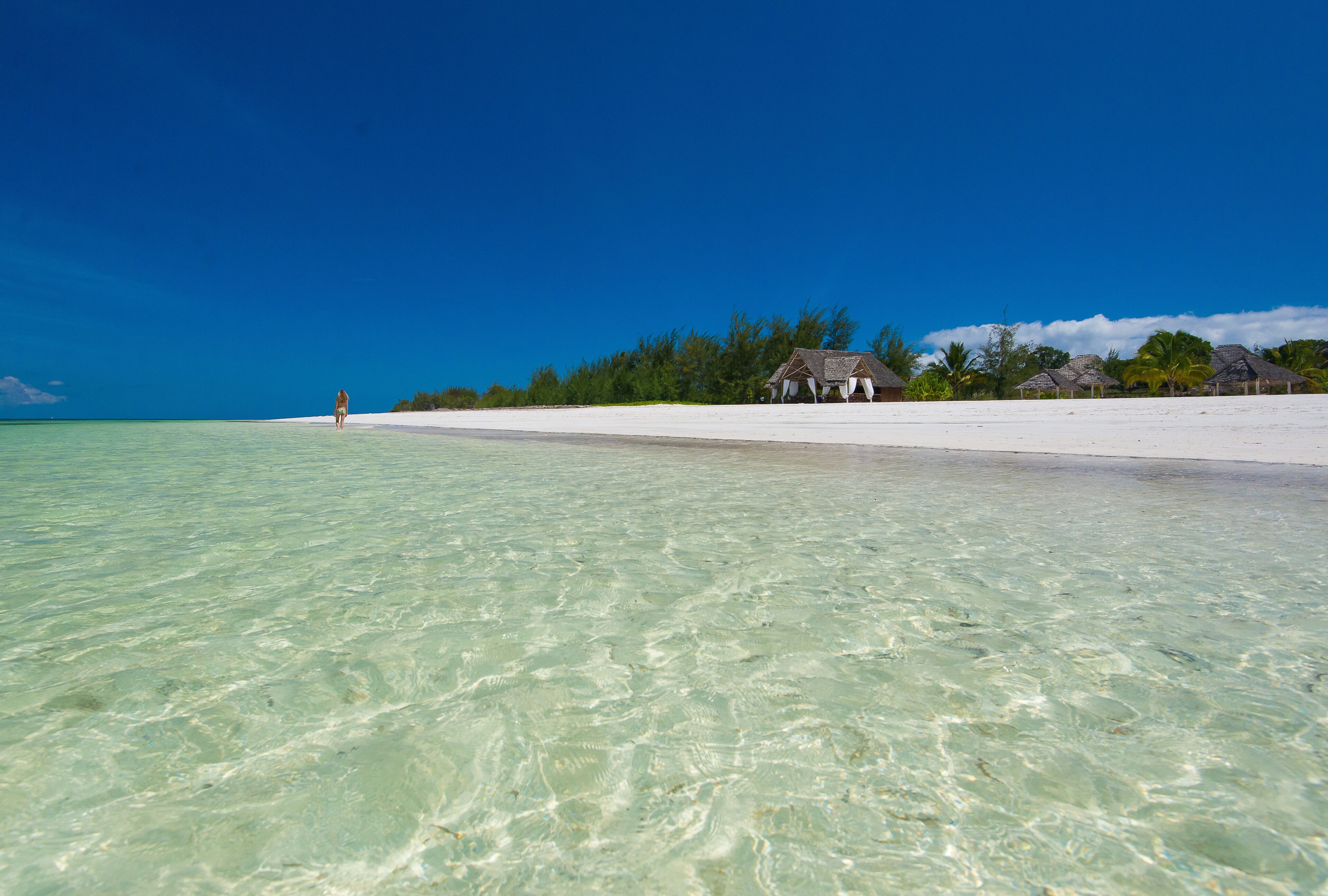 Una playa con agua turquesa cristalina, una persona caminando y cabañas de techo de paja al fondo.