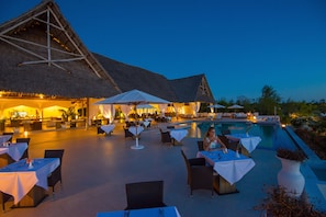 An outdoor dining area with tables and chairs, a pool, and a thatched-roof building.