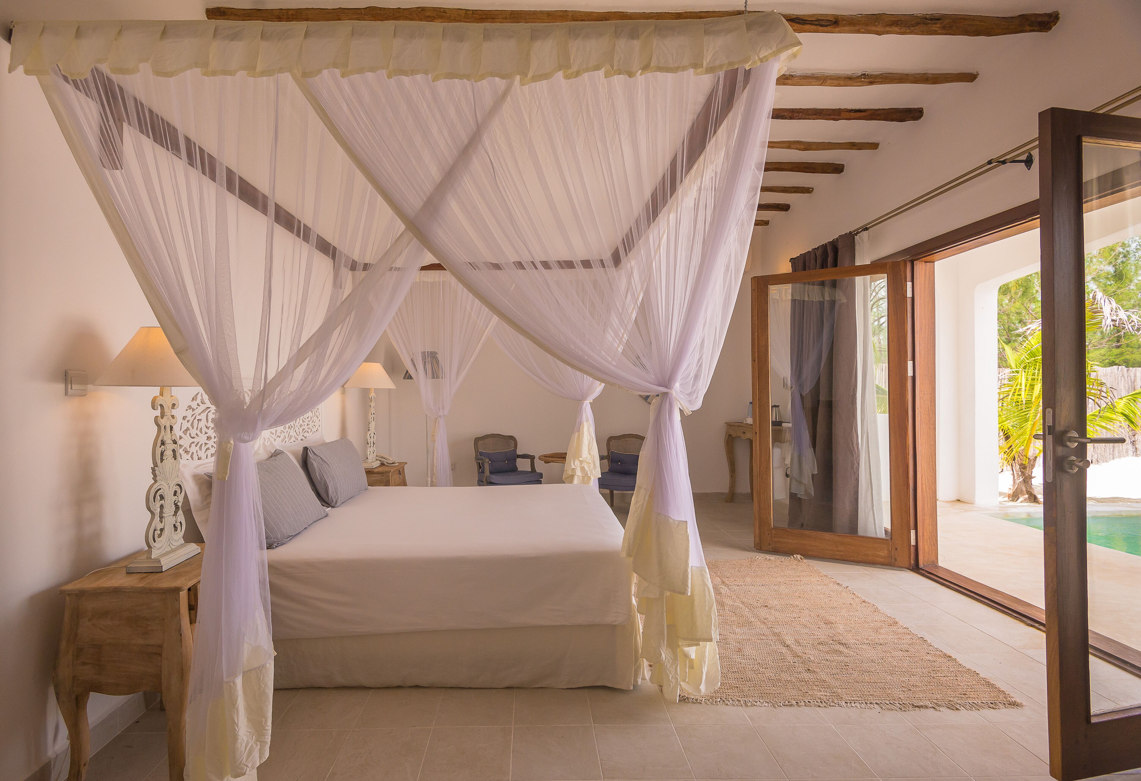 A bedroom with a four-poster bed, a nightstand, a lamp, and a view of a swimming pool through a glass door.