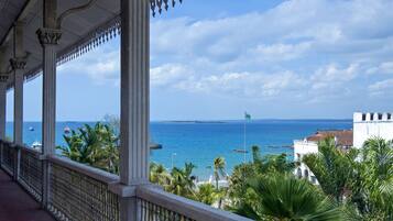 A balcony with a view of the sea, palm trees, and a white building with a red roof.