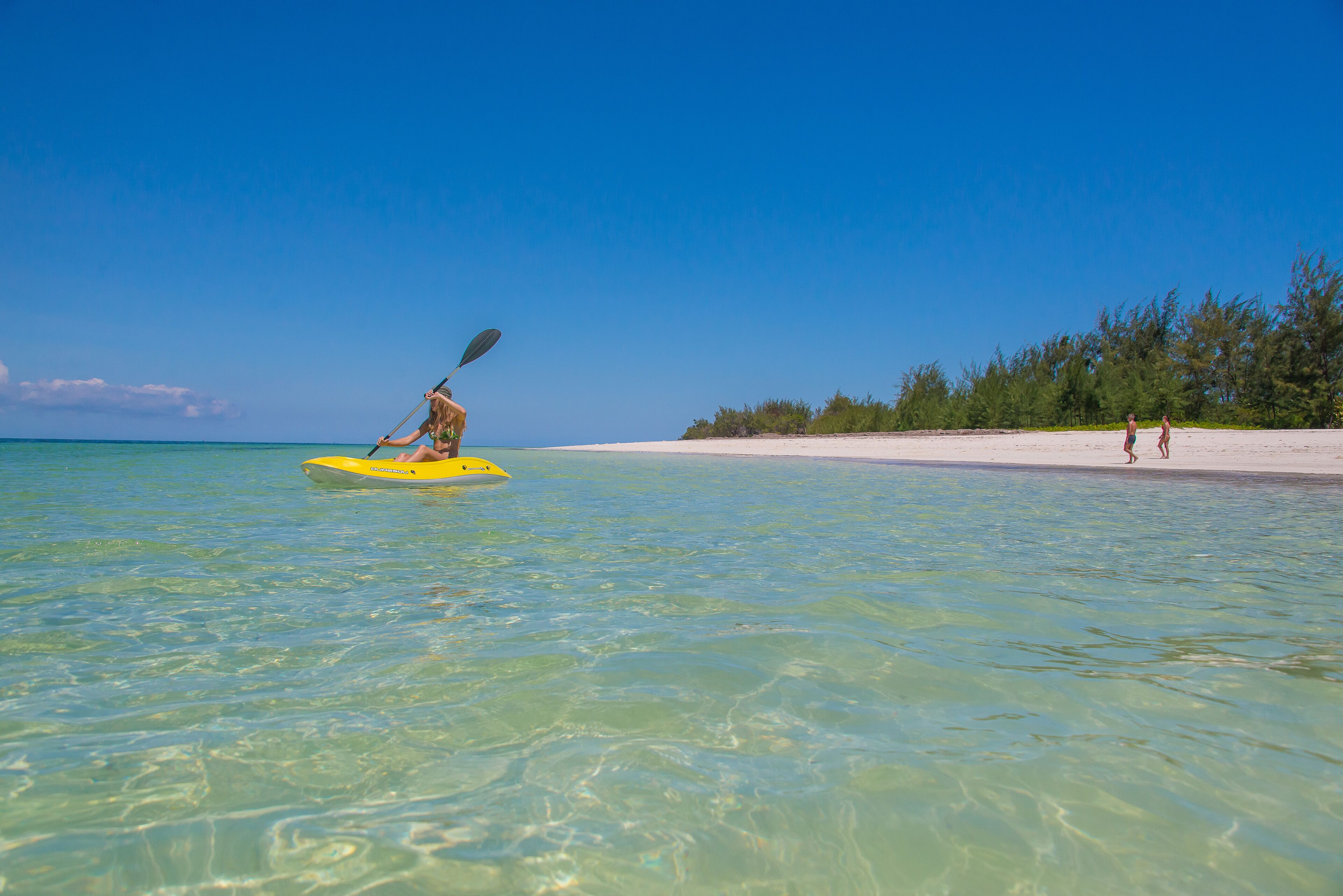 Une plage aux eaux claires, avec un kayak sur l’eau et une rive sablonneuse bordée d’arbres.