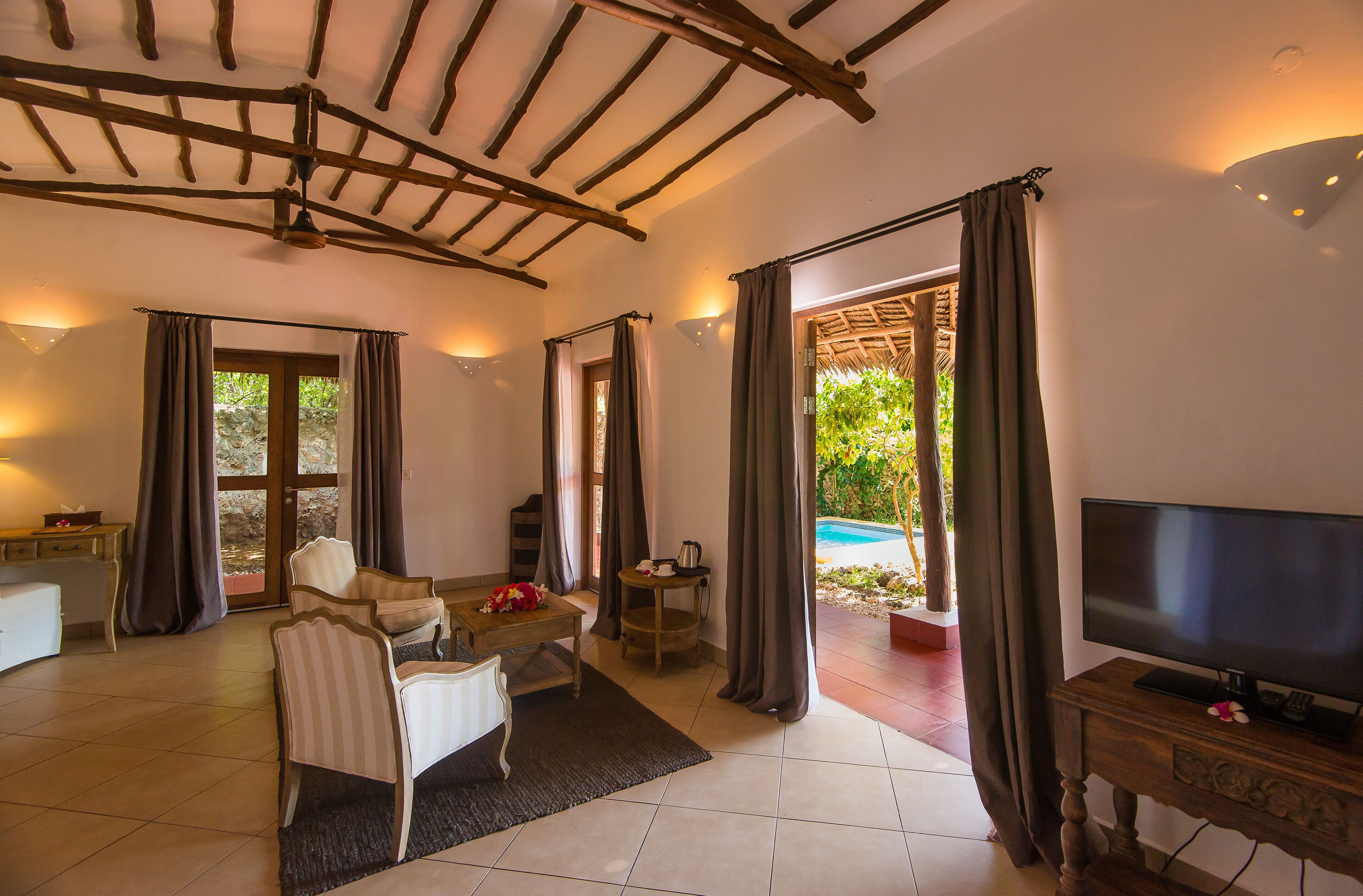 Habitación con televisión, una mesa de comedor con sillas, una mesita con teléfono y hervidor, y vista a una piscina al aire libre.