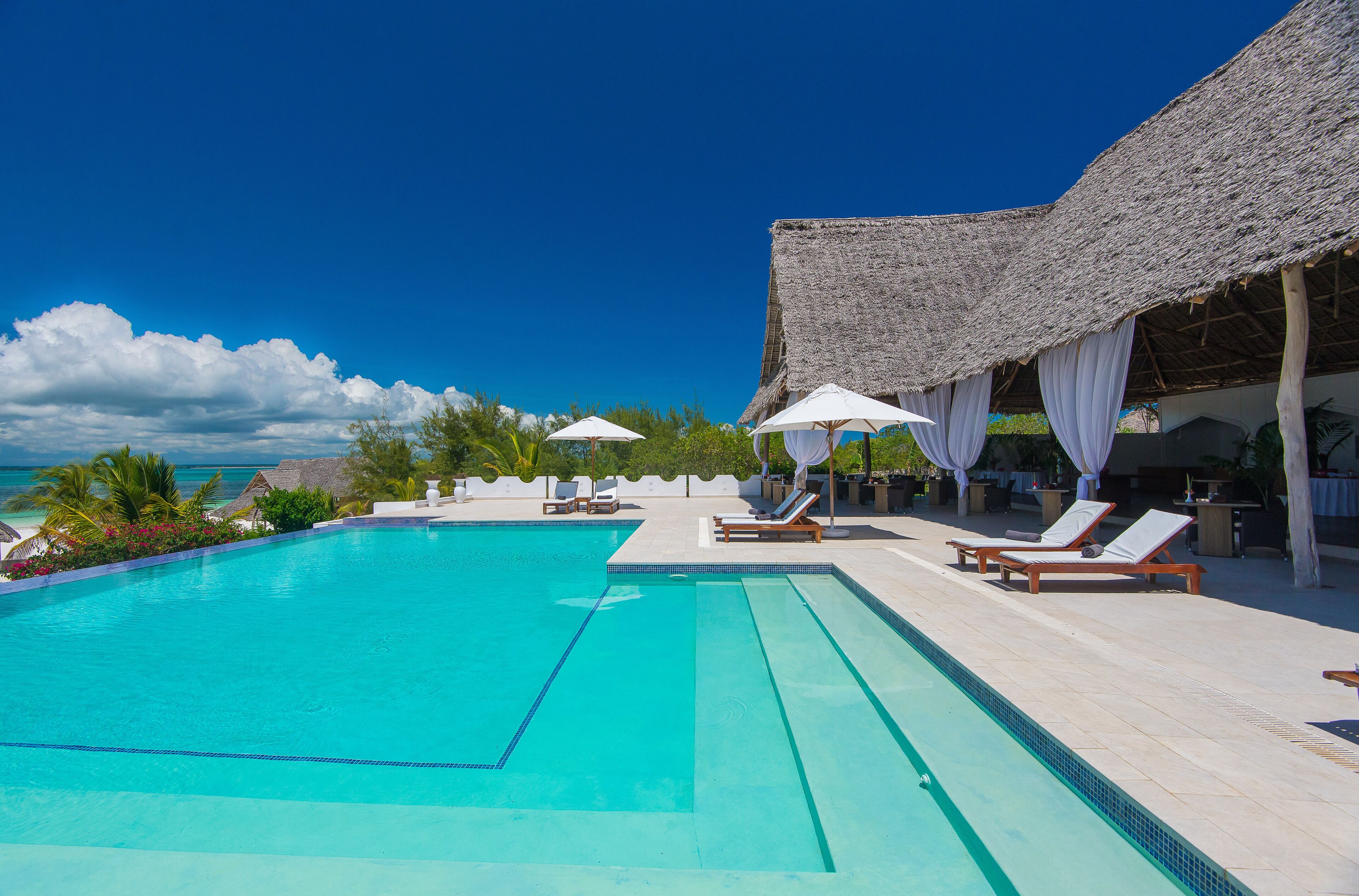 A clear blue swimming pool with lounge chairs and umbrellas, surrounded by a thatched-roof structure and palm trees.