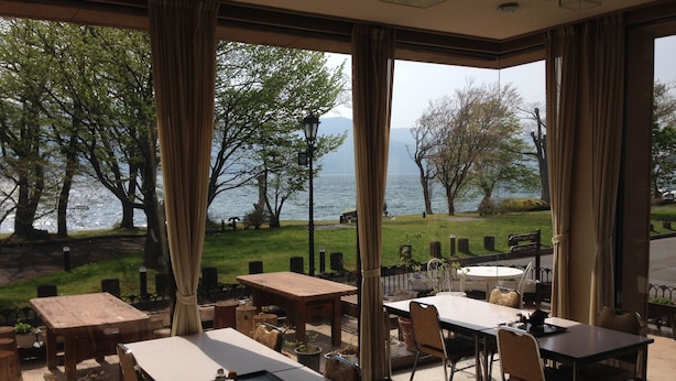 Un restaurante con terraza frente a un lago, con mesas y sillas de madera, y amplias ventanas con cortinas.