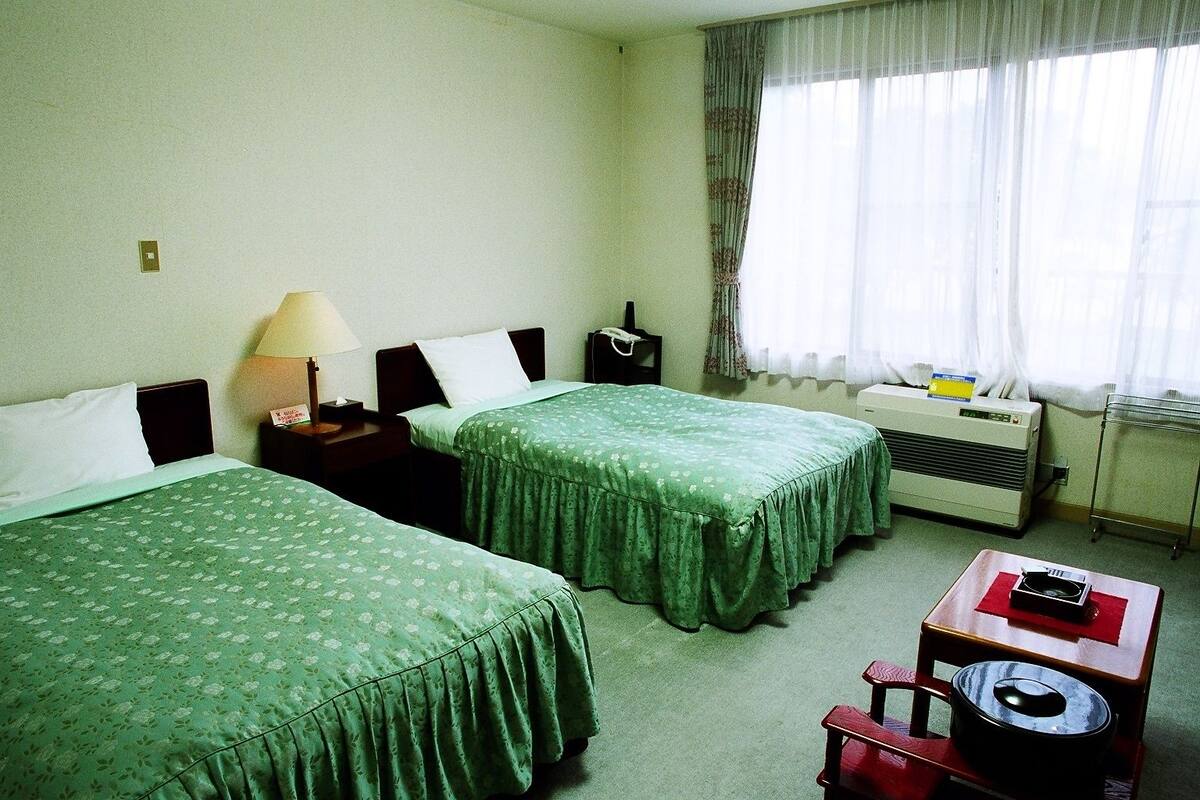 Ein Hotelzimmer mit zwei Einzelbetten, einem Nachttisch mit Lampe, einem Heizkörper, einem kleinen Tisch mit Wasserkocher und einem Fenster mit Vorhängen.