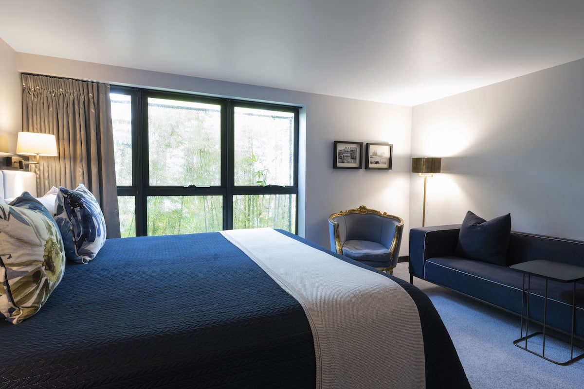 Une chambre d’hôtel avec un grand lit, un canapé, un fauteuil et une petite table. Il y a deux fenêtres avec des rideaux, une lampe et des tableaux encadrés au mur.