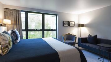 Habitación de hotel con una cama grande, un sofá, una silla y una mesita pequeña. Hay dos ventanas con cortinas, una lámpara y cuadros en la pared.