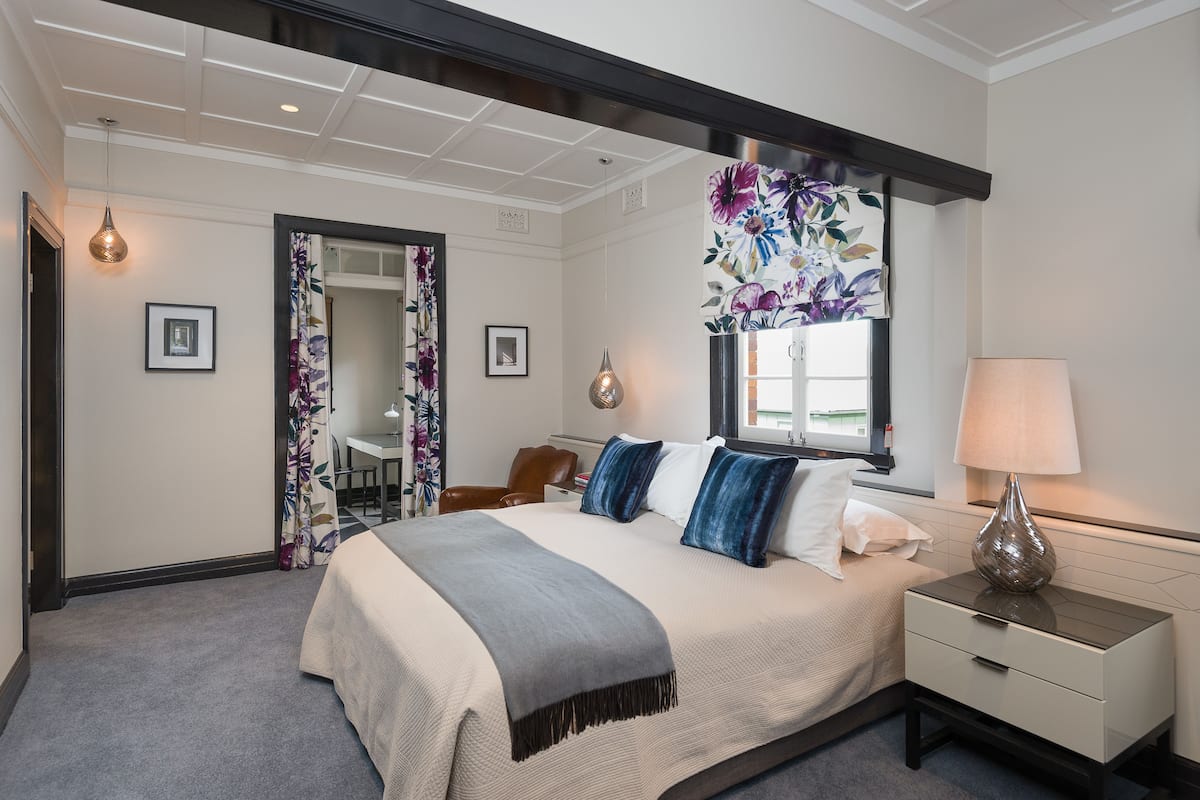 Une chambre d’hôtel avec un lit, une table de chevet, une lampe et une fenêtre ornée d’un papier peint à motifs floraux.