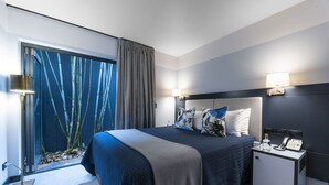 Habitación de hotel moderna con una cama grande, una mesita de noche con lámpara y vistas a bambú a través de un ventanal amplio.