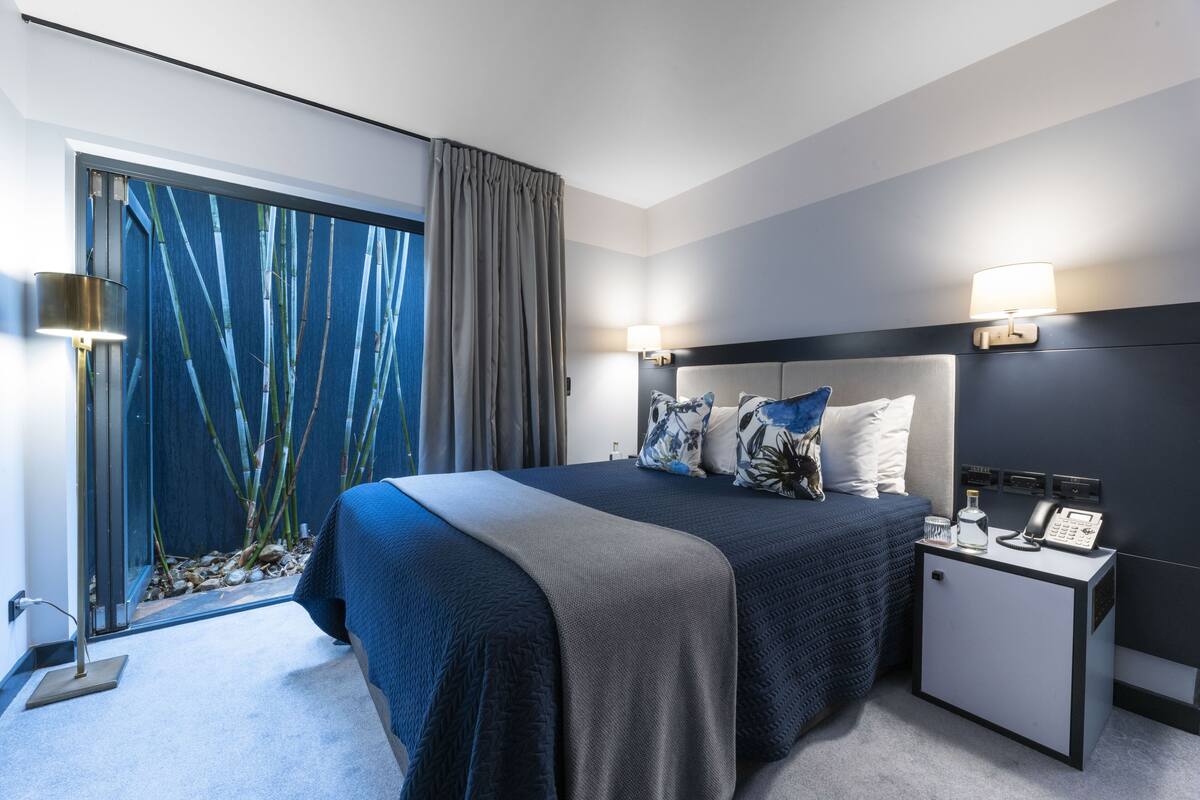 Une chambre d’hôtel moderne avec un grand lit, une table de chevet avec une lampe et une vue sur des bambous, visible à travers une grande fenêtre.