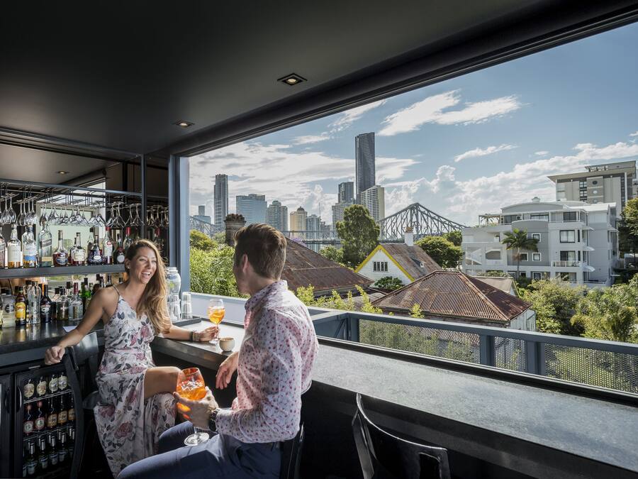A bar with a city view, shelves with bottles, and a person serving drinks.