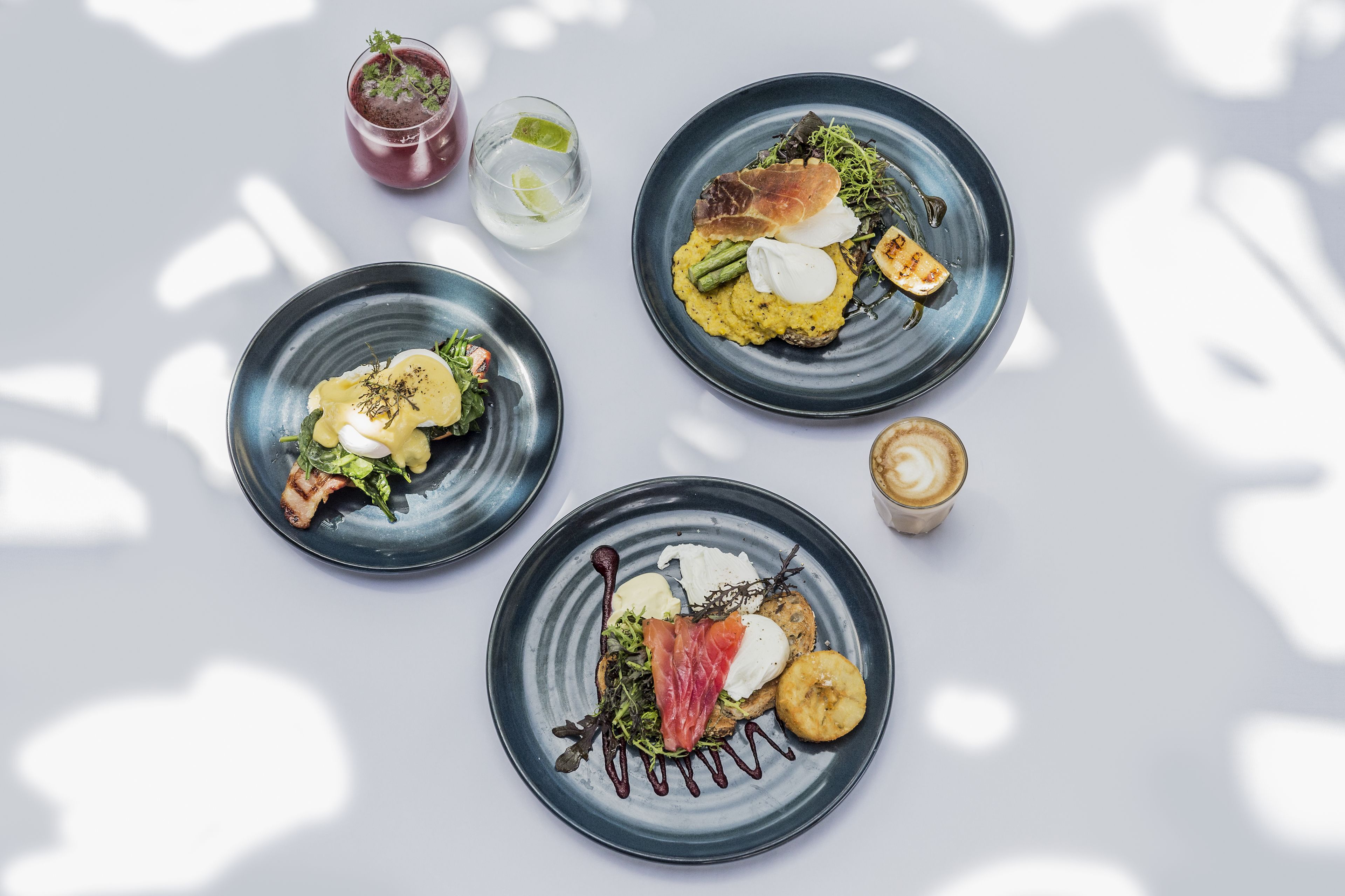 Three plates of food with various items including greens, meats, and sauces, and two drinks on a white surface.